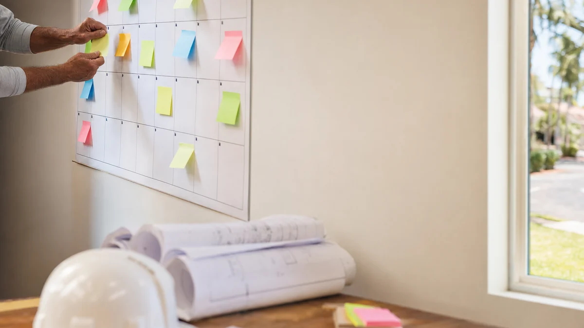 Pool builder pinning blank notes to a crowded build calendar next to rolled pool plans