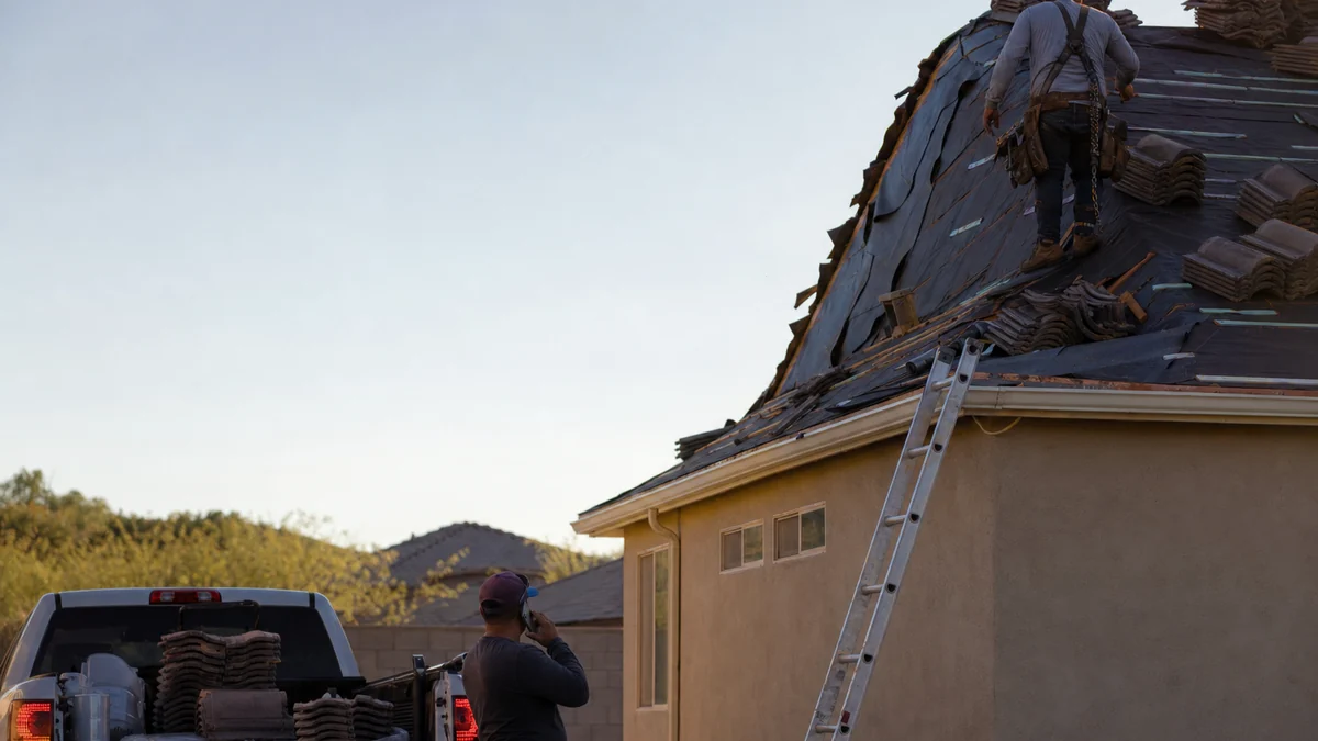Roofer working on a roof while a teammate on the ground returns a customer's call from the truck